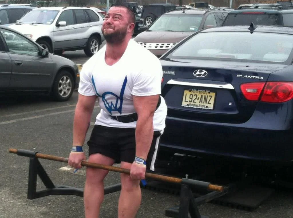 A man with a beard, wearing a white T-shirt and black shorts, is lifting a heavy metal barbell in a parking lot. There are multiple parked cars behind him, including a blue Hyundai Elantra with a yellow license plate. The man appears to be straining as he lifts.