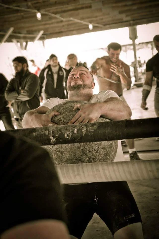 Man in a white shirt holding a large rock in an arm wrestling match, surrounded by other men, some smiling and one shirtless, under a wooden shelter.