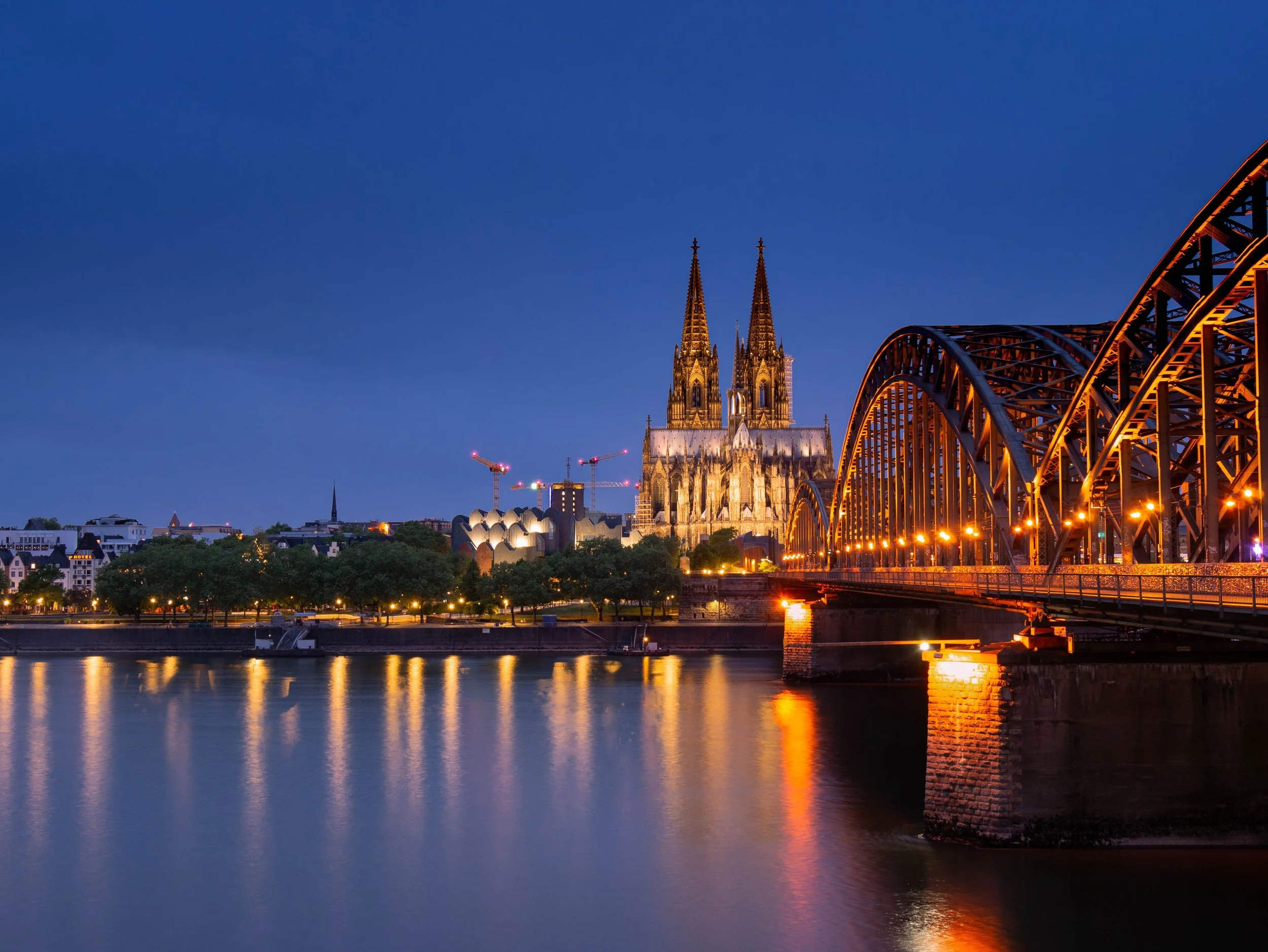 Night view of a river with illuminated cityscape, including a Gothic cathedral and a bridge, under a dark blue sky.