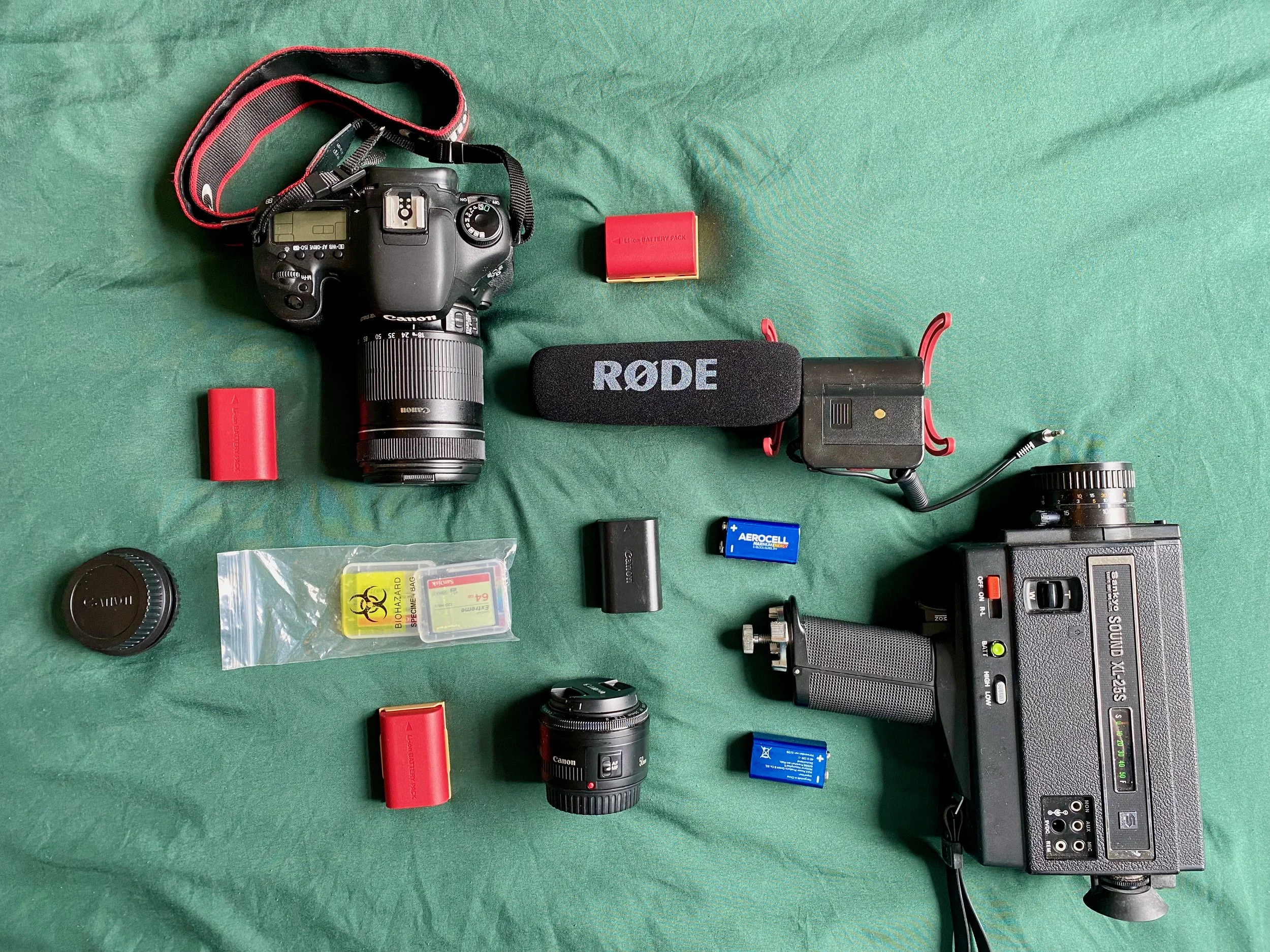A collection of camera equipment on a green surface, including a DSLR camera, a lens, a Rode microphone, batteries, a lens cap, and various cables and accessories.