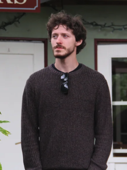 Young man with curly dark hair and a beard, wearing a brown sweater and sunglasses hanging from the collar, standing outdoors in front of a building with windows and a door.