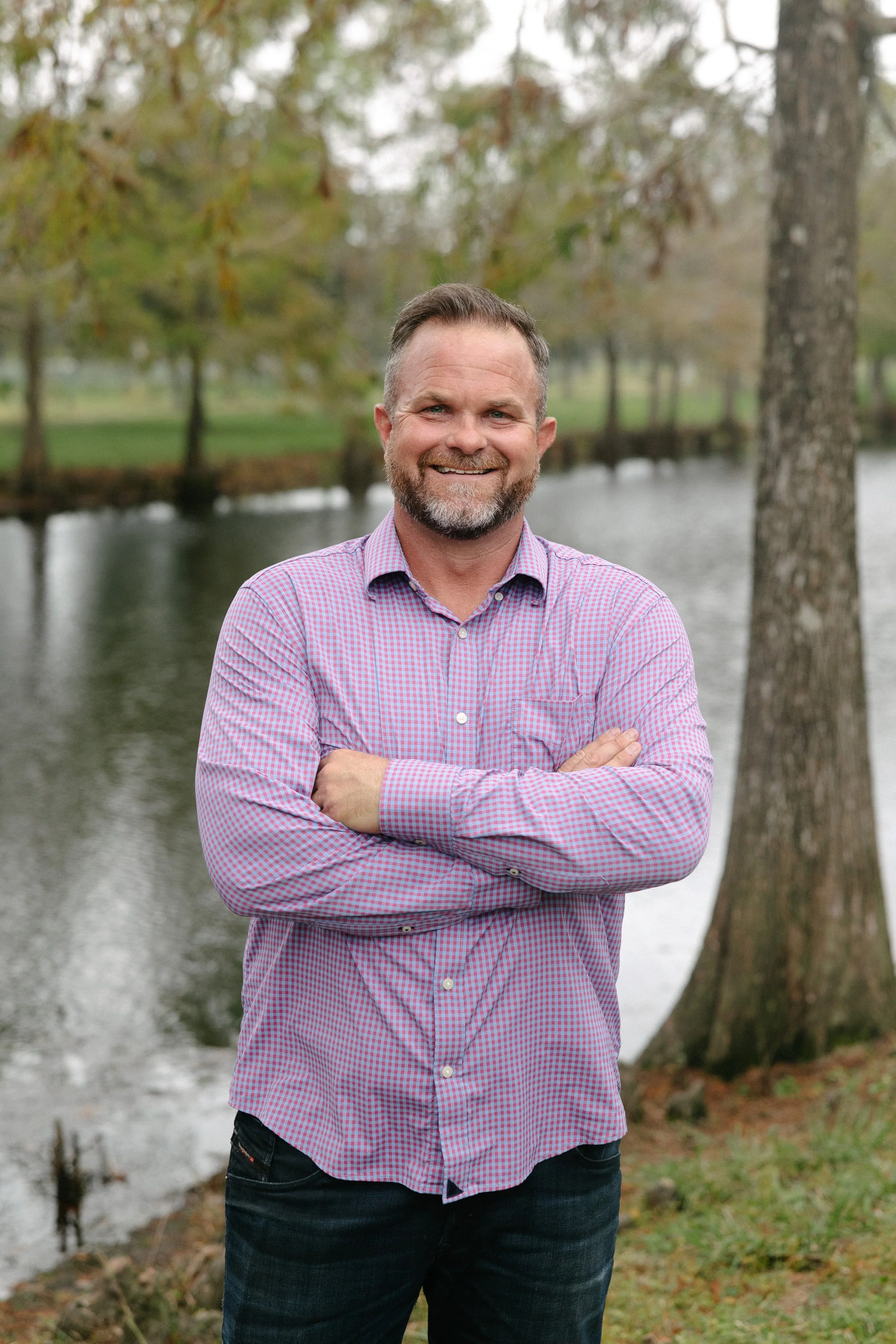 A man with a beard and short hair, wearing a pink checkered shirt and dark jeans, standing outdoors by a river with trees in the background.
