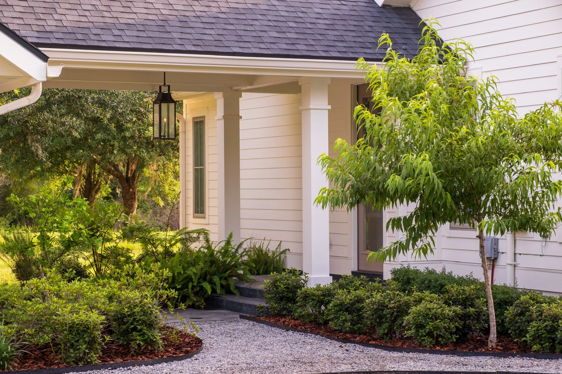 Front porch of a white house with dark shingles, surrounded by lush green trees and shrubs, gravel pathway, and a tree on the right.