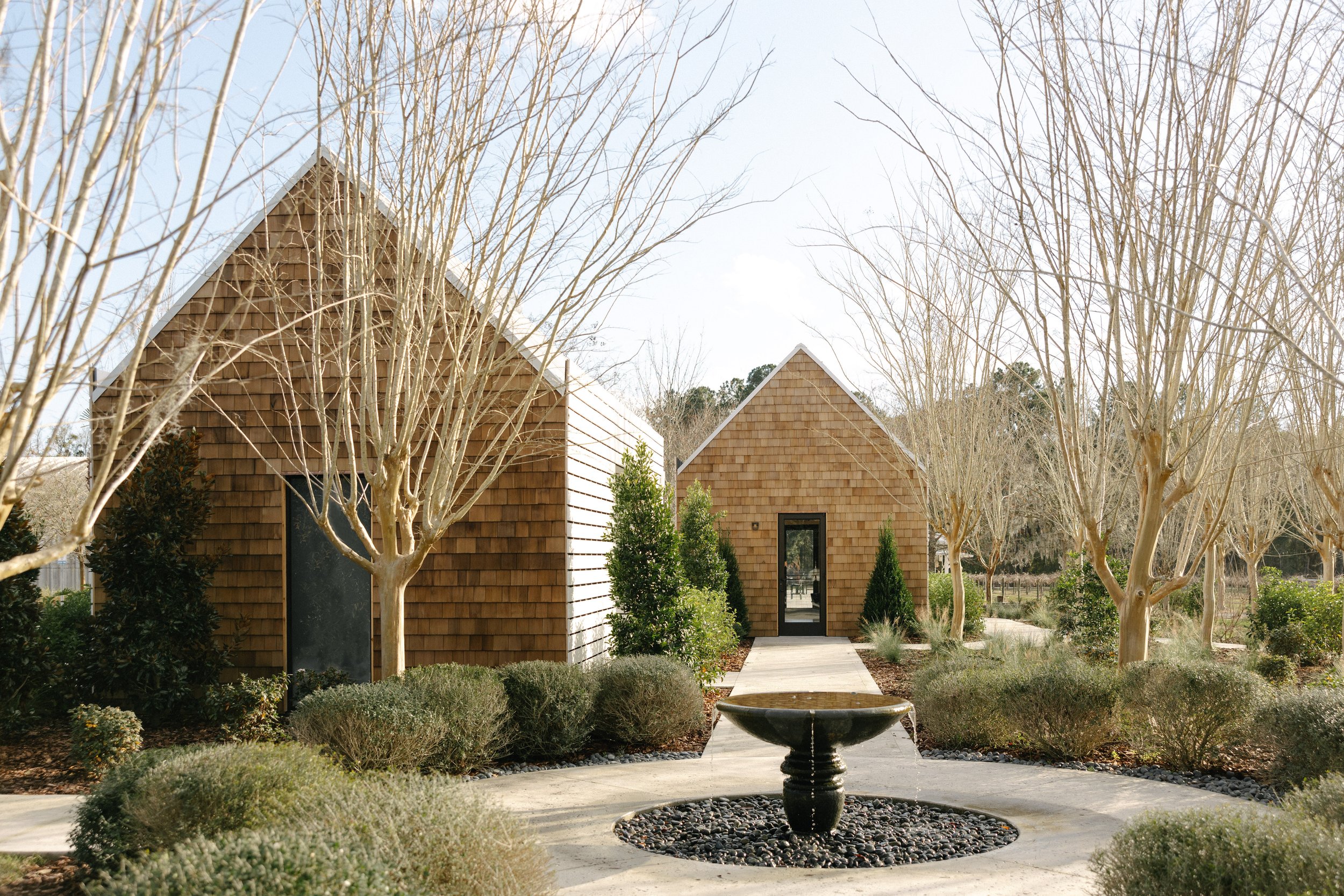 A modern house with wooden siding, surrounded by leafless trees and shrubs, with a pathway leading to the entrance and a central fountain in the garden.