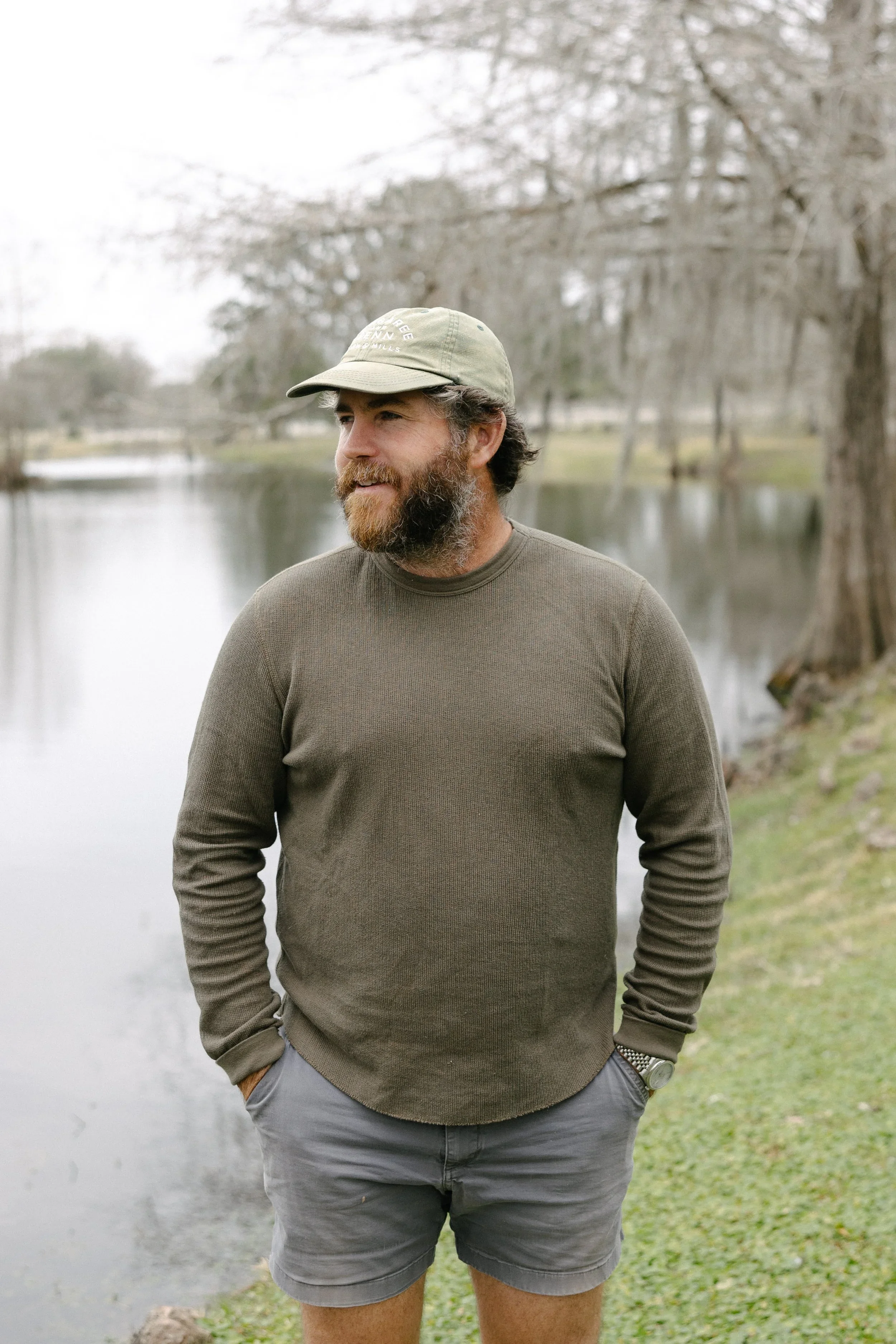 A man with a beard wearing a light green baseball cap, a brown long-sleeve shirt, and gray shorts standing outdoors near a body of water with leafless trees in the background.