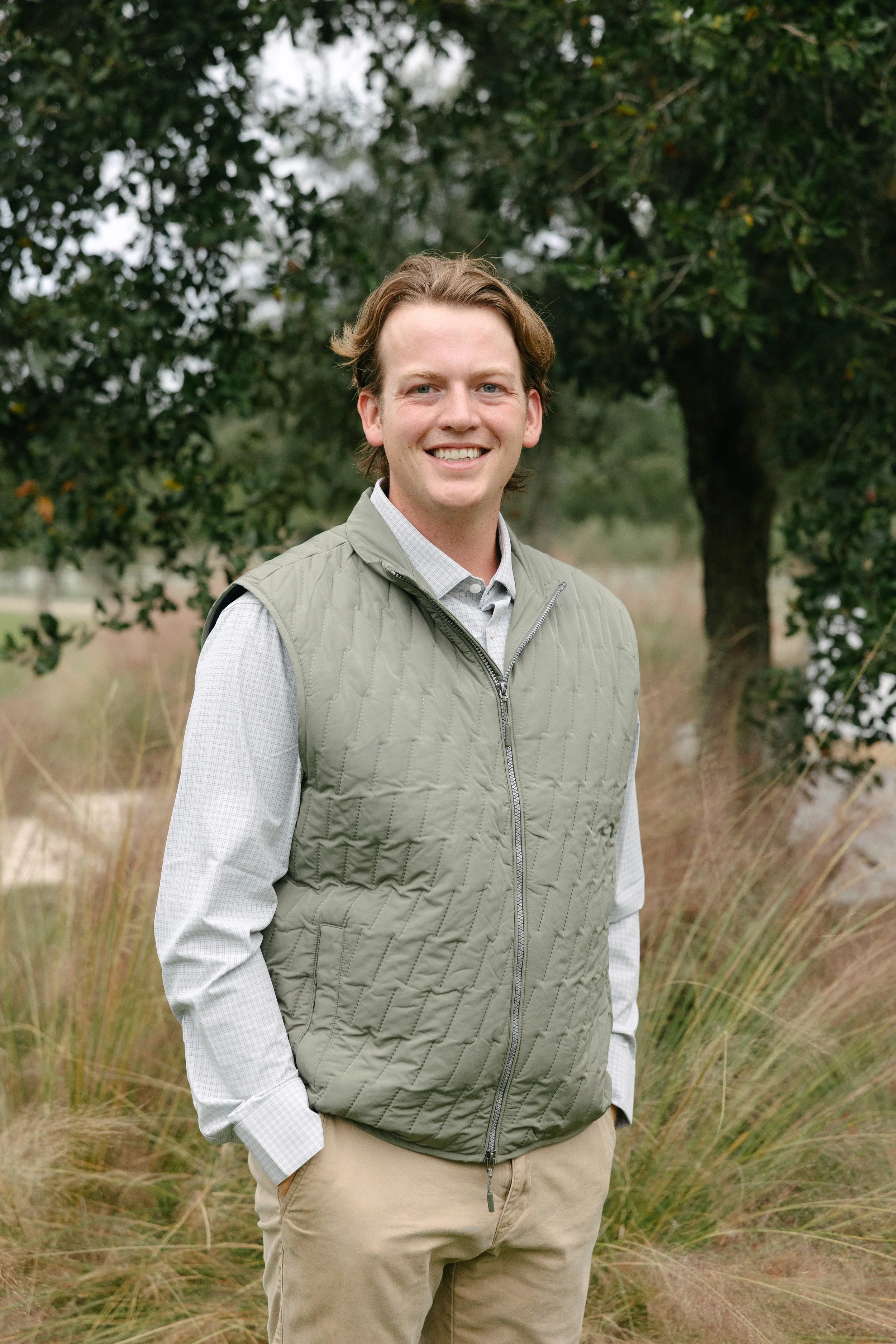 A smiling man standing outdoors in front of a tree and grass, wearing a light green vest over a gray checkered shirt and khaki pants.