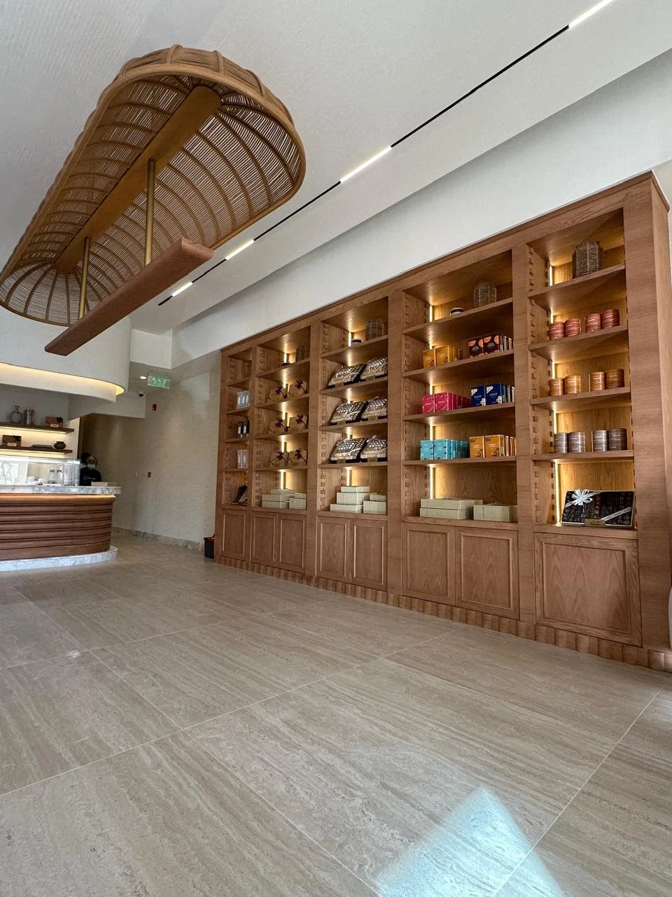 Interior of a modern retail store or cafe with wooden shelving displaying various items, a curved counter on the left, and a wicker light fixture hanging from the ceiling.