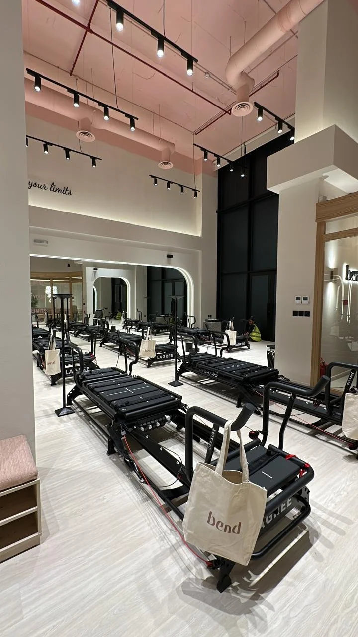 Fitness studio with multiple reformer Pilates machines, some with tote bags hanging on the side, in a well-lit room with a high ceiling and modern decor.