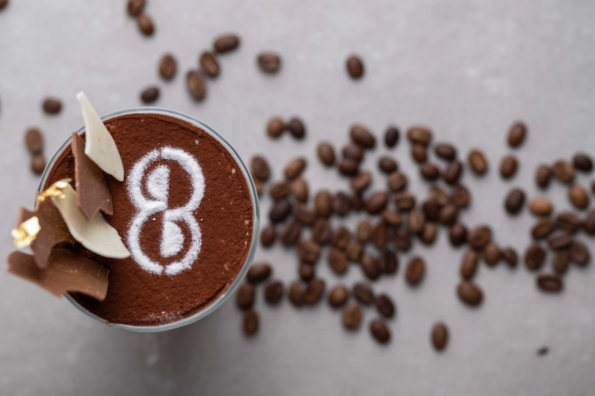 Close-up of a cup of tiramisu topped with cocoa powder, white chocolate shavings, and a sugar design, surrounded by scattered coffee beans on a light surface.