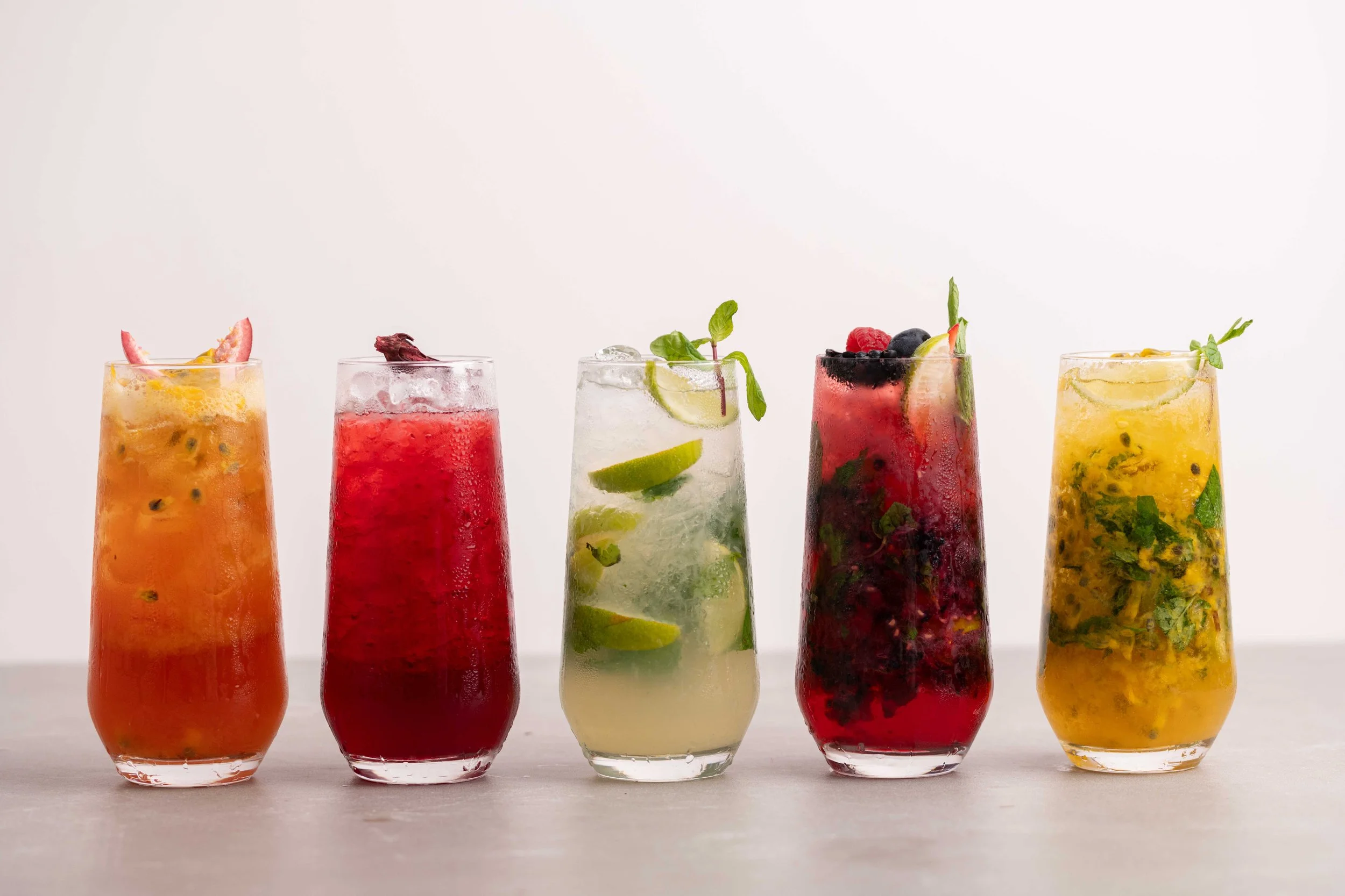 Five colorful fruit drinks in tall glasses with various fruit garnishes, on a white surface.