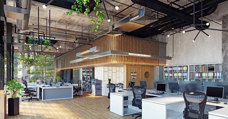 Open-concept modern office space with white desks and black ergonomic chairs, featuring a wood-paneled ceiling installation, large windows, plants, and colorful bookshelf walls.