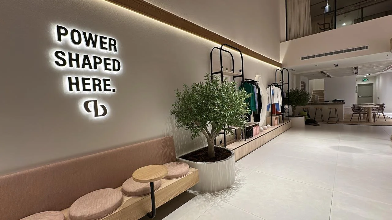 Modern interior of a retail store or lounge area with a lit sign that reads 'Power Shaped Here' and a logo below. There are clothing racks, a large potted plant, and seating with cushions.