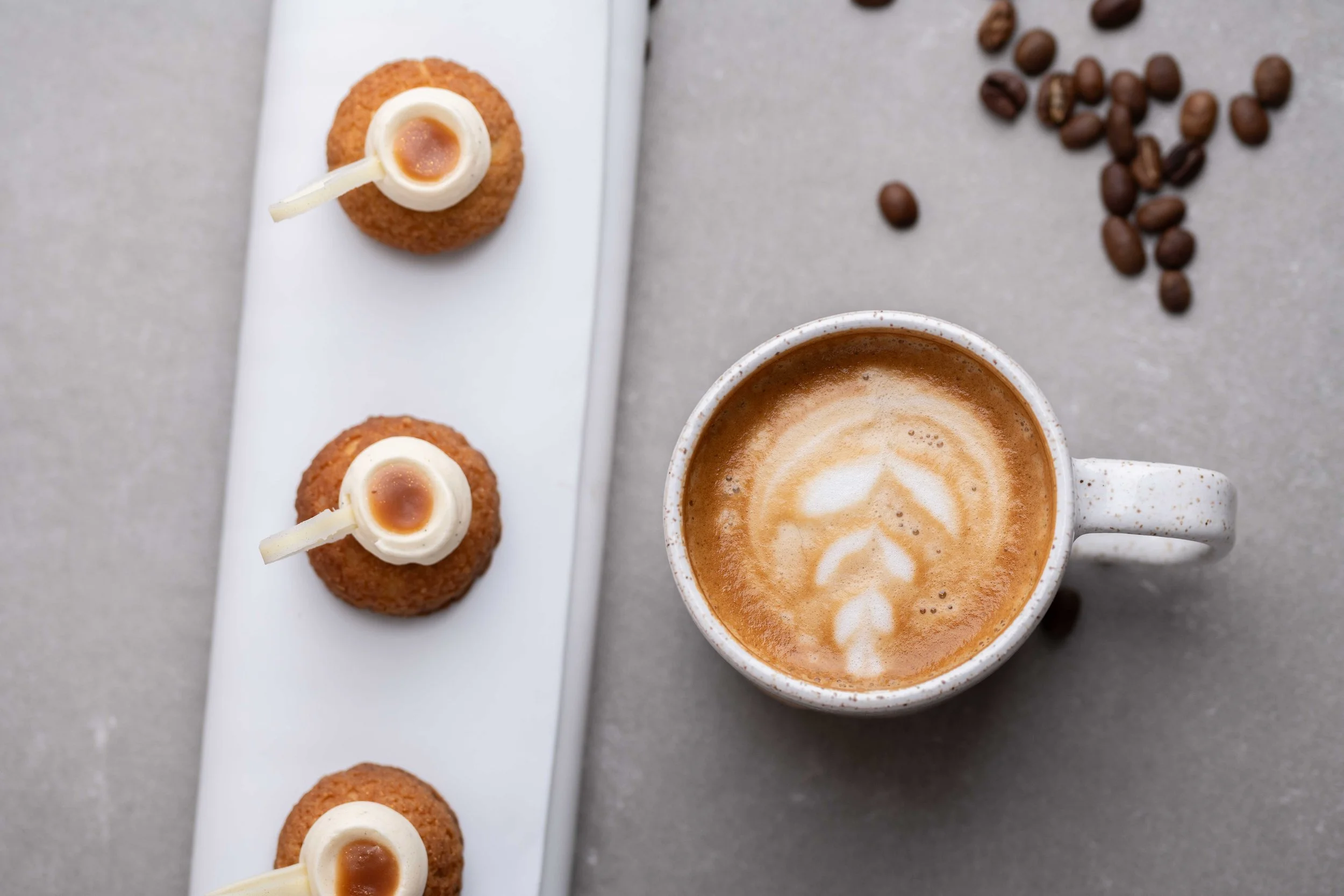 A white tray with three small round fried snacks topped with white sauce and a brown dot, a cup of coffee with foam art, and scattered coffee beans on a gray surface.