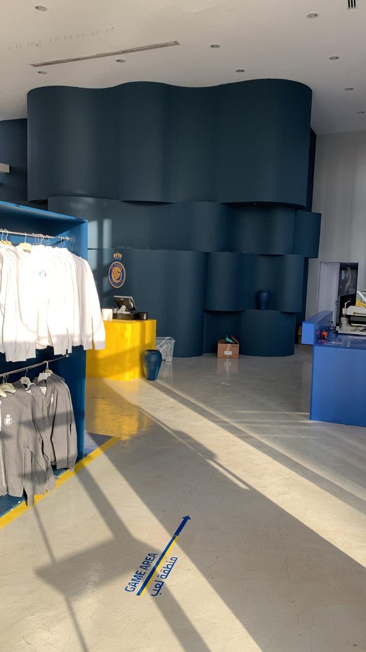 Interior of a modern store with clothing on the left, a yellow checkout counter with a display, and a blue sculpture or wall feature in the background. Sunlight casts shadows on the white floor, which has a blue and yellow sign indicating the 'Game Area.'