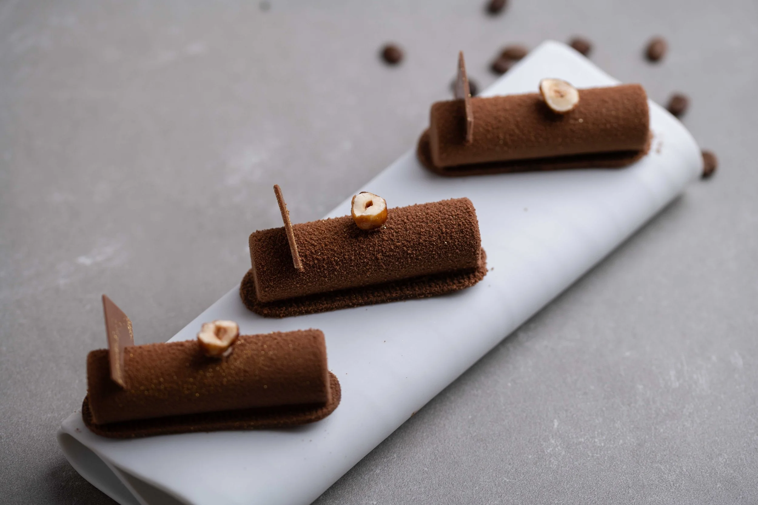 Close-up of a row of elegant chocolate desserts with hazelnuts on top, placed on a white serving platter on a gray surface.