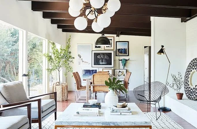 Living room with white walls, large windows, and modern decor. Includes a white sofa, a modern black wire chair, wooden dining table with six chairs, and various framed artwork on the wall. A large white vase with green leaves on the coffee table, and a cluster of globe pendant lights hanging from a dark wooden ceiling.