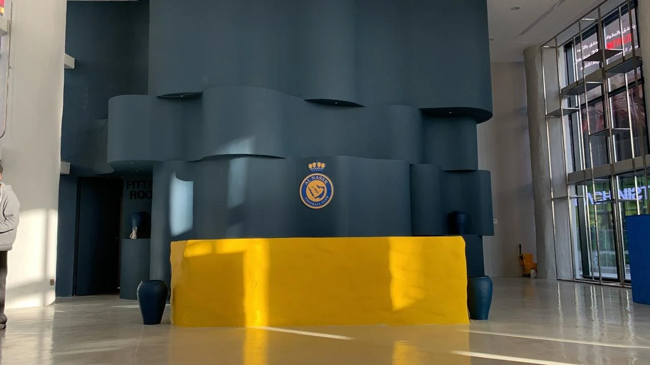 Indoor lobby area with a dark blue wall featuring the Nassau Soccer Club logo, and a large yellow rectangular structure in front. There are glass doors and windows on the right side, allowing sunlight to enter and cast shadows on the floor.