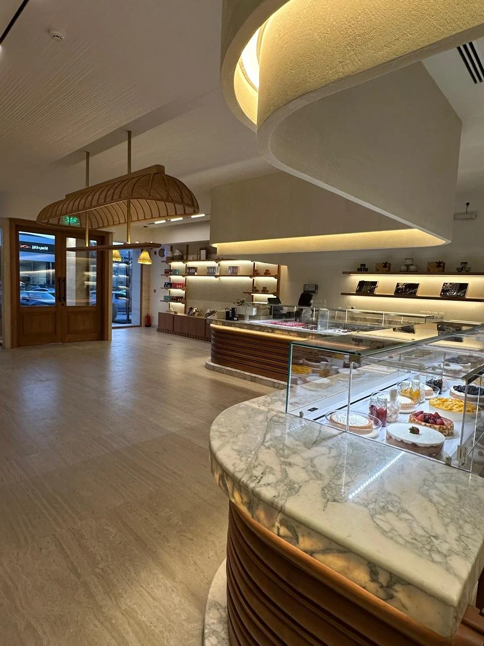 Interior of a modern bakery or cafe with marble display counter featuring pastries and desserts, wooden accents, decorative shelving, and a curved ceiling design.