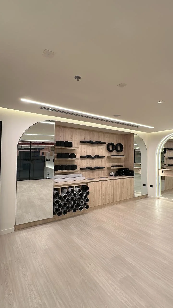 Empty fitness studio with wooden wall shelves holding black dumbbells, yoga mats, and resistance bands. Rolled-up yoga mats stored in a cubby, large mirrored wall, and light wood flooring.