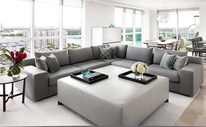 Modern living room with gray sectional sofa, white ottoman, and large windows showing city view, with a dining area and hanging chair in background.
