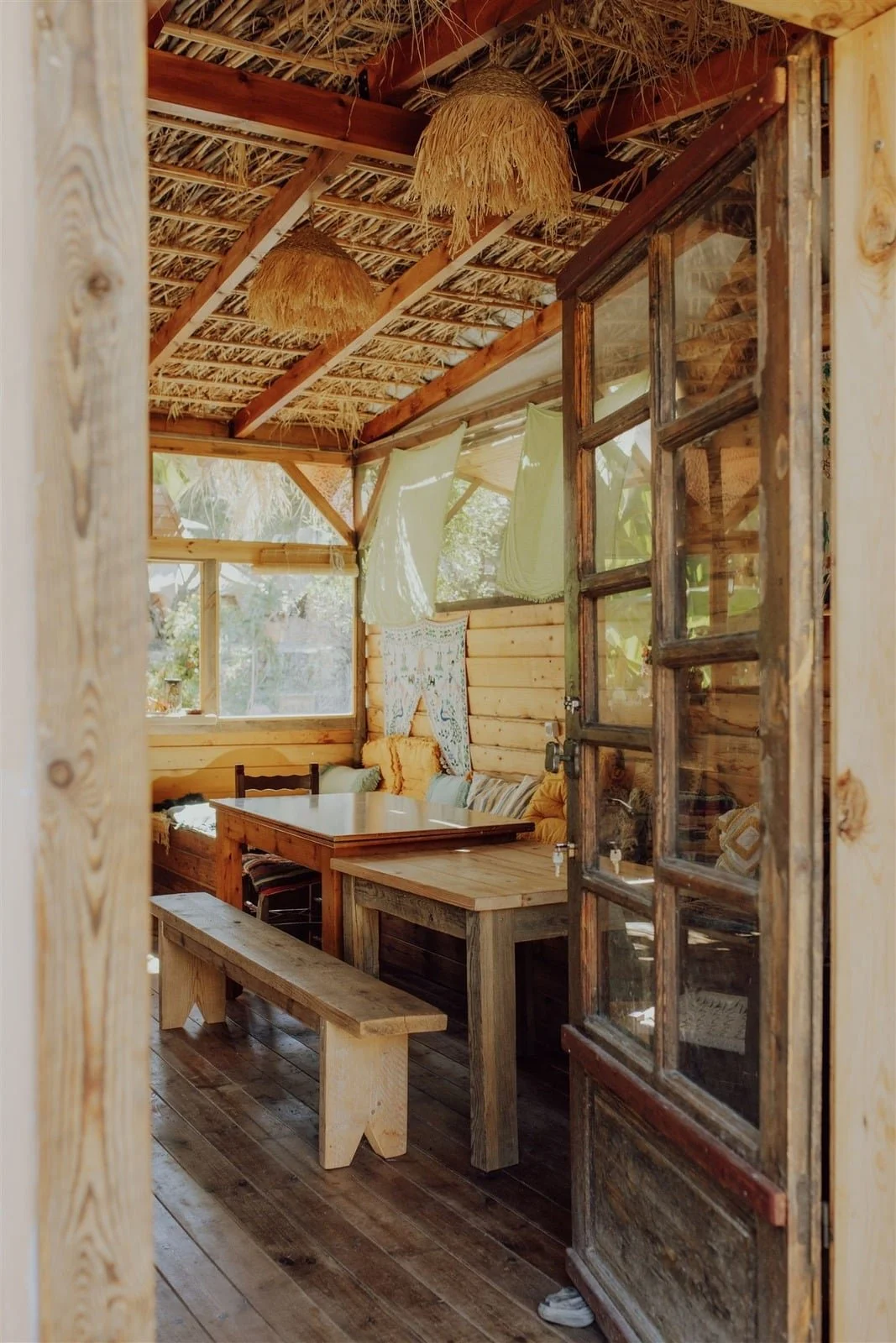 Inneneinrichtung eines rustikalen Holzhauses mit einem Tisch, Bänken, Sitzkissen und gestickten Decken, Blick durch eine alte Holztür.