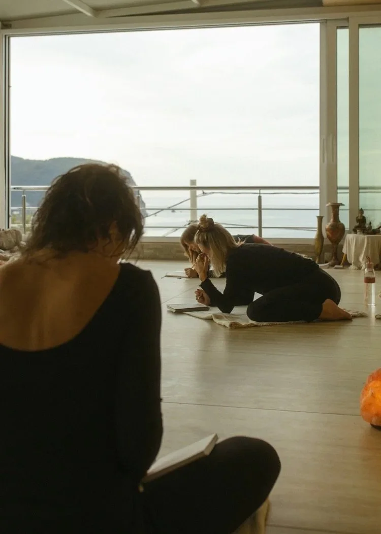 Menschen in einer Yoga- oder Meditationsklasse, die auf dem Boden auf Matten sitzen und in einer ruhigen, hellen Umgebung mit großen Fenstern blicken, die eine Aussicht auf Wasser und Hügel zeigen.