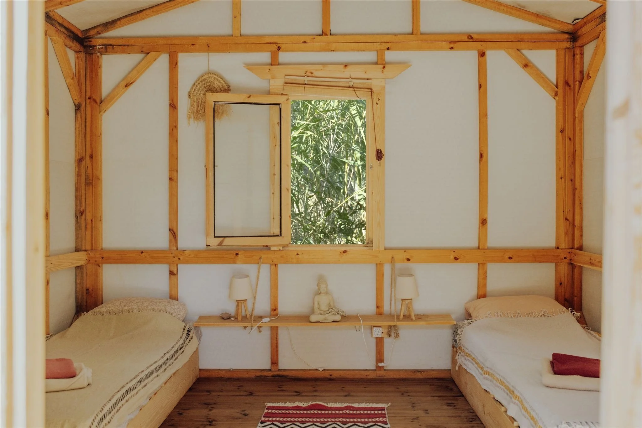 Ein Schlafzimmer mit zwei Betten, Holzwänden, einem kleinen Fenster in der Mitte, einer kleinen Statue und Lampen auf einem Regal, ein Teppich auf dem Holzboden.