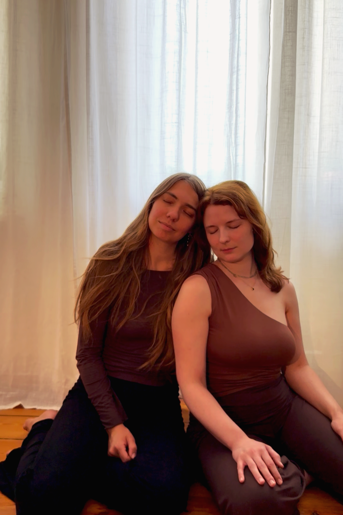 Zwei Frauen sitzen nebeneinander vor einem hellen Vorhang, ihre Augen sind geschlossen, beim Entspannen oder Meditieren.