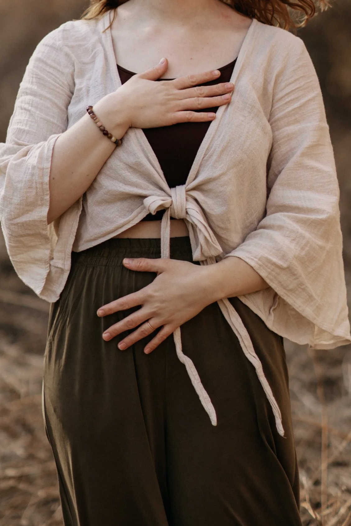 Eine Frau trägt ein schwarzes Top, ein beigefarbenes Jacke und braune lockere Hosen, hält eine Hand auf die Brust und die andere auf den Bauch.