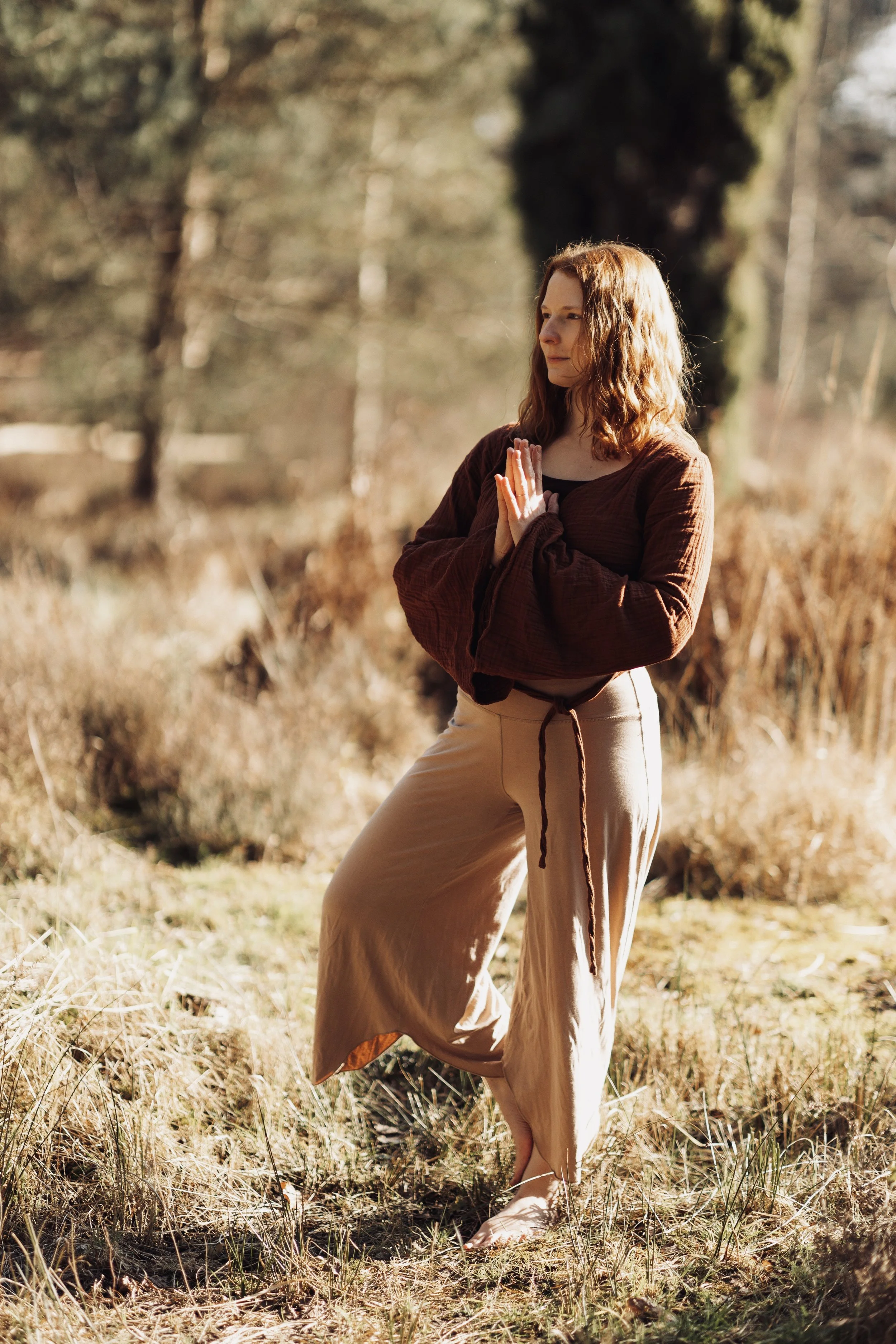 Eine junge Frau praktiziert Yoga im Freien während des Sonnenaufgangs oder Sonnenuntergangs, umgeben von Natur mit Bäumen und trockenem Gras.