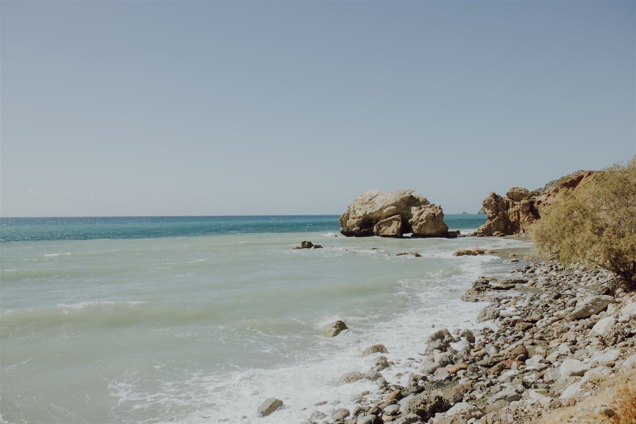 Ein Strand mit Felsen und grünlich-blauem Wasser unter einem klaren blauen Himmel.