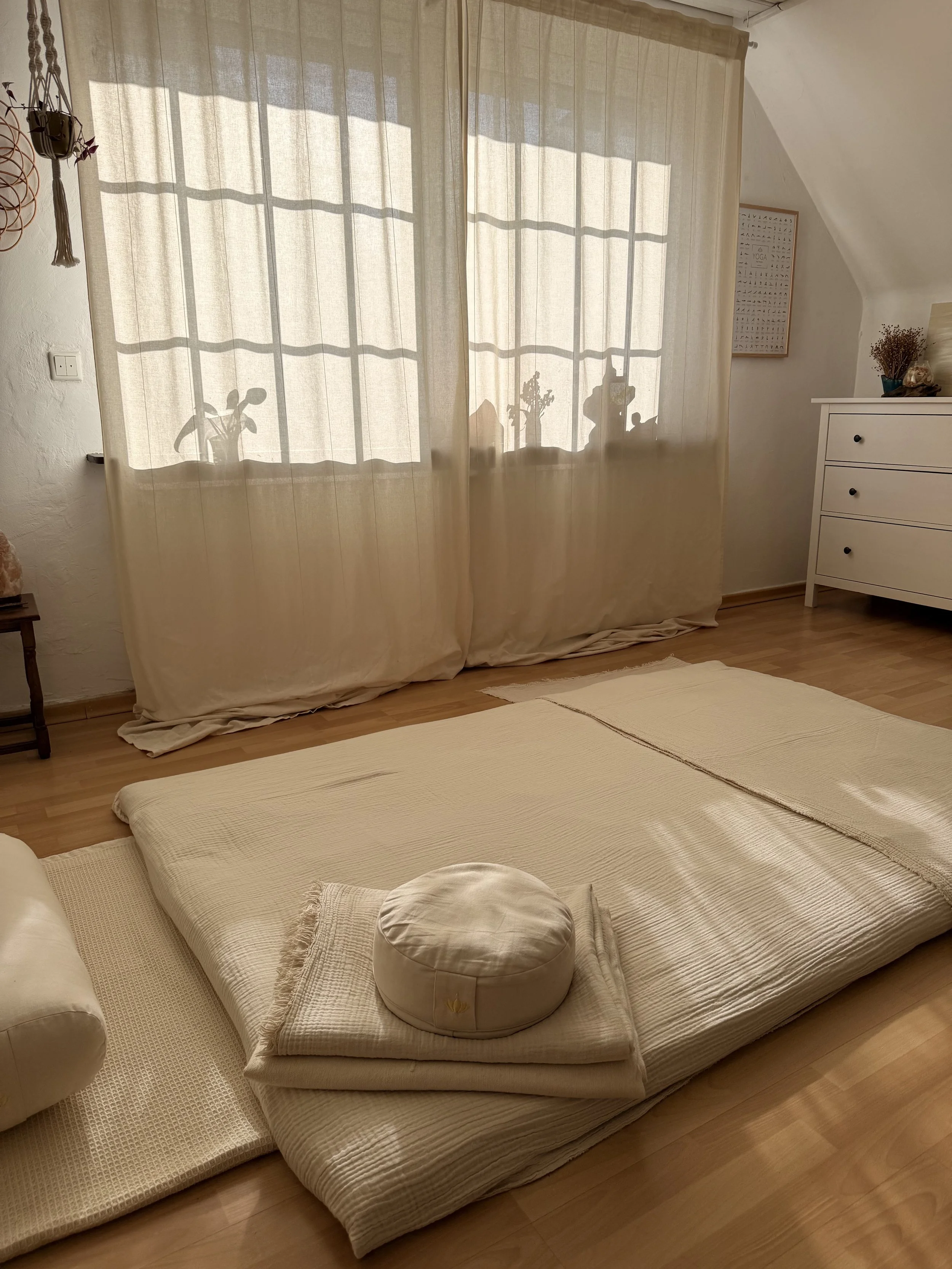 Ein Schlafzimmer mit einem beige Bett, eines runden Kissen, einem Teppich, Vorhängen, die Schatten von Pflanzen an die Wand werfen, und einem weißen Nachttisch mit Dekorationen.