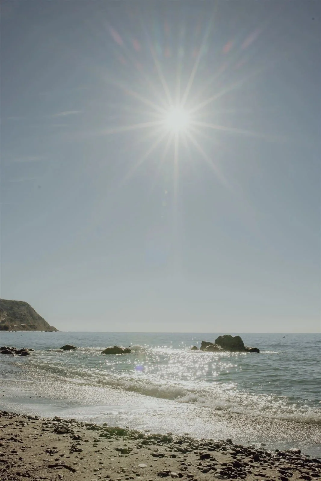 Sonnenstrahlen scheinen auf das Meer, Blick auf einen Strand mit Steinen und Felsen, klare Himmel.