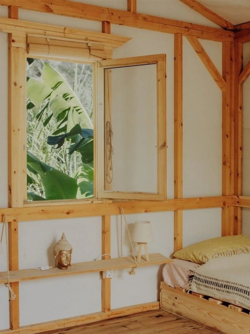 Ein Schlafzimmer mit Holzrahmen und einem offenen Fenster, das einen Blick auf grüne Pflanzen zeigt. Es gibt eine Bettrahmen mit Bettzeug, eine Nachttischlampe, eine Statue eines Buddha-Kopfes und ein kleines Regal.