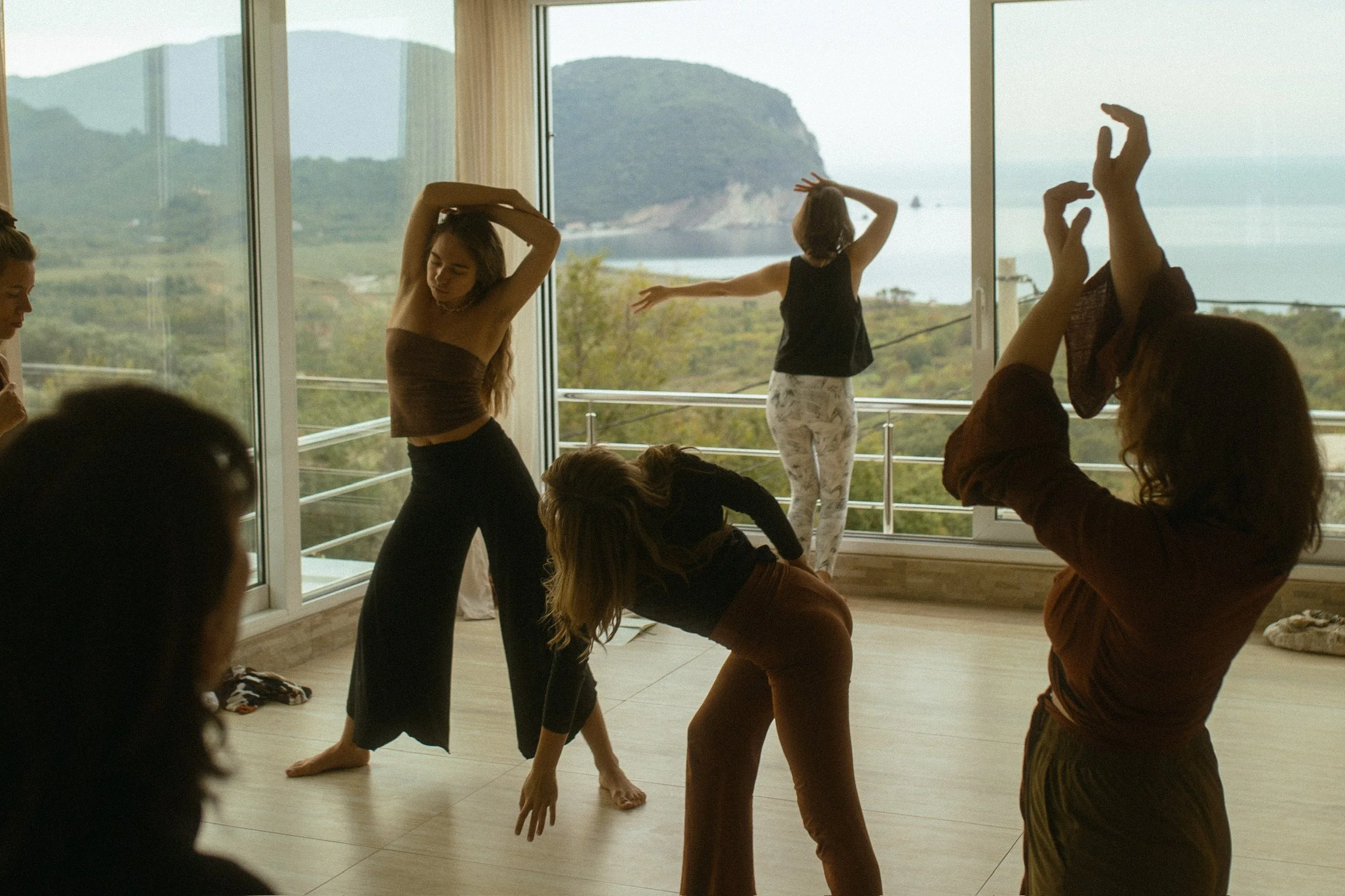 Menschen, die Yoga und Tanzübungen in einem hellen Raum mit großen Fenstern und Blick auf eine grüne Landschaft praktizieren.
