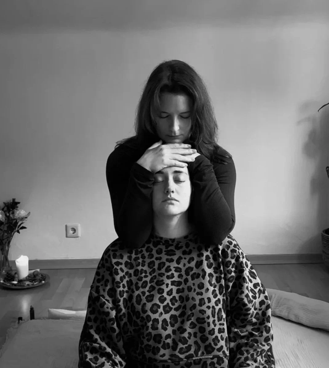 Zwei Frauen, eine sitzend mit geschlossenen Augen und Leopardenmuster-Kleidung, die andere stehend mit den Händen auf ihrem Kopf und der Kopfseite, in einem Raum mit einfacher Einrichtung und Pflanzen.