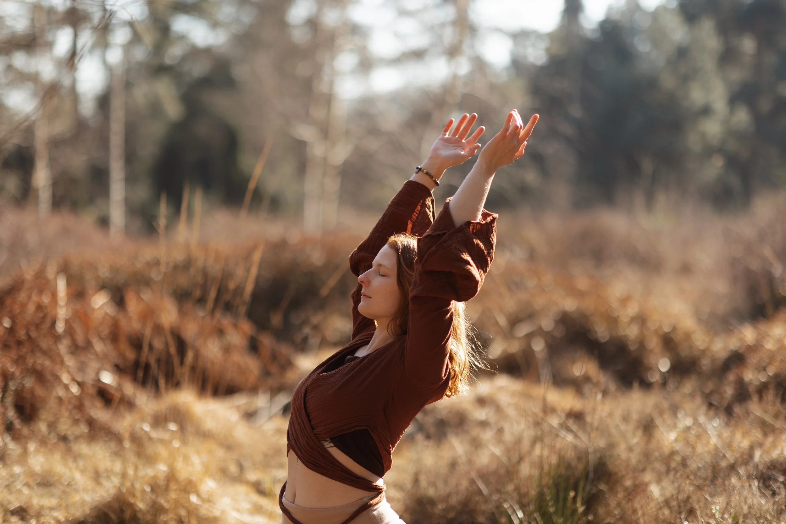 Eine Frau steht im Freien in einem Wald, hebt die Arme hoch und schließt die Augen in einer entspannten Haltung während des Sonnenuntergangs.