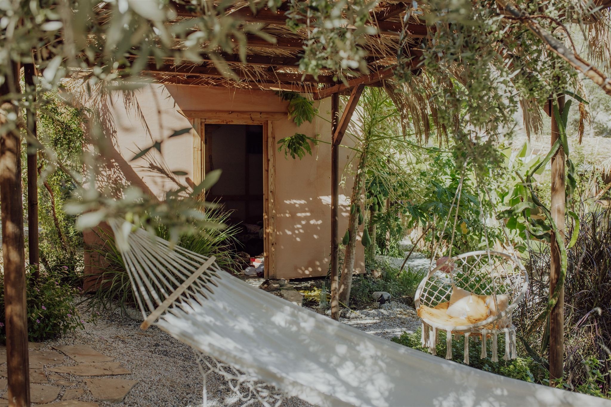 Ein kleines Hüttchen in einem Garten, umgeben von grünen Pflanzen, Bäumen und Sonnenschein, mit entspannter Atmosphäre.