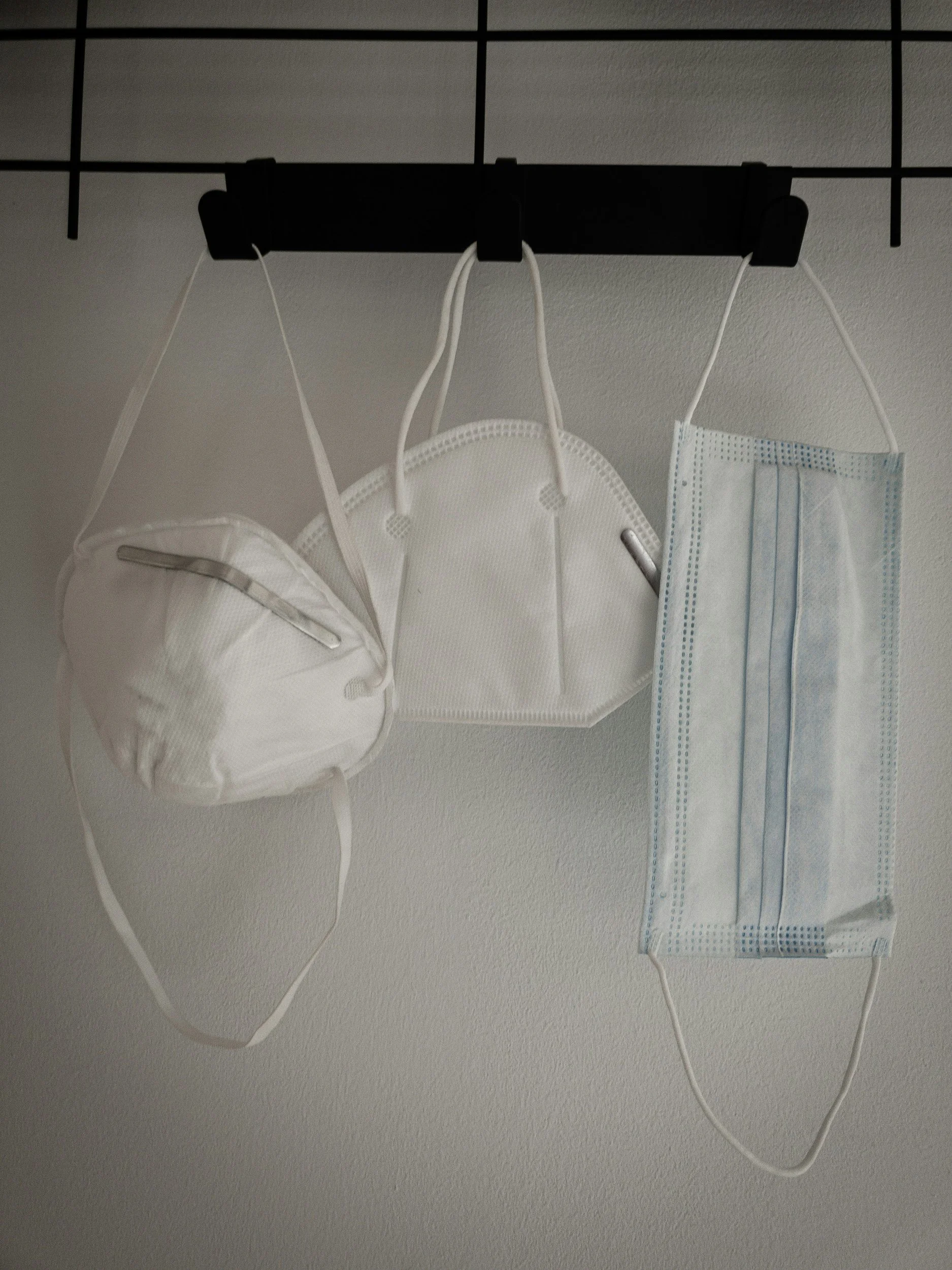 Three face masks hanging from a black hook on a ceiling grid, including a white N95 mask, and two disposable surgical masks, one slightly folded.