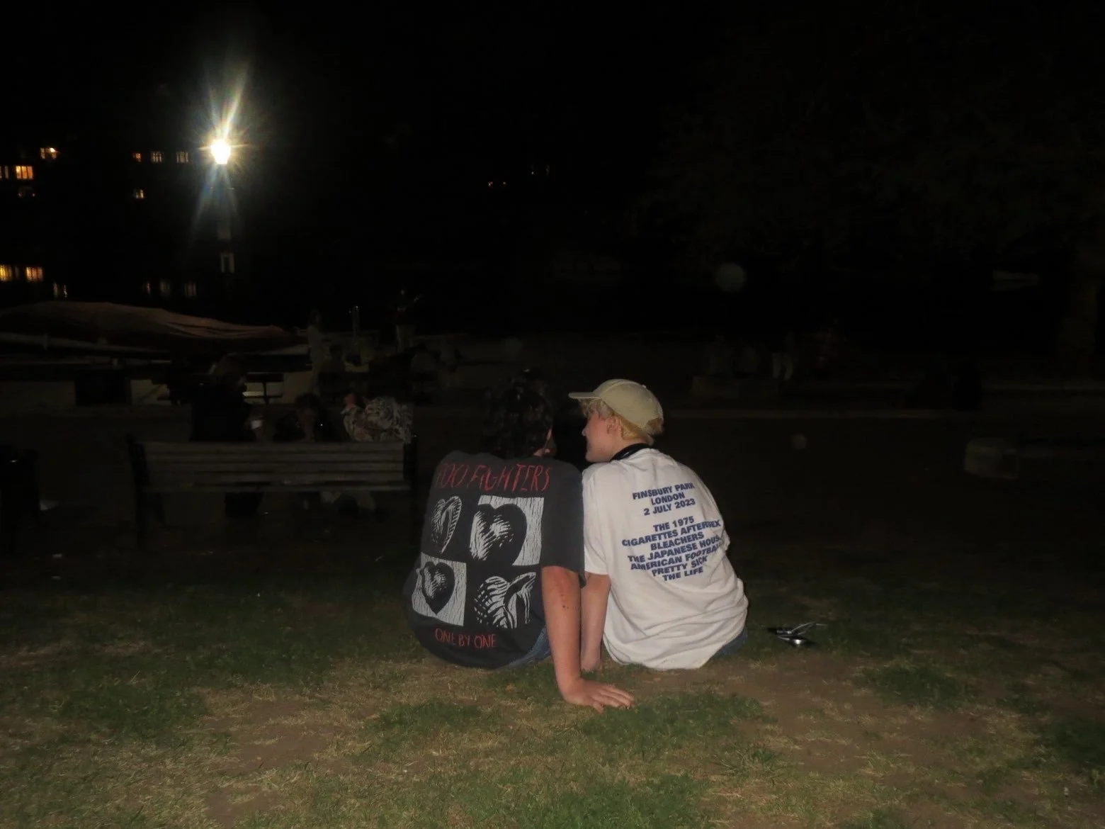 Two young people sitting together on grass in a park at night, with their backs to the camera, sharing a quiet moment. One wears a black shirt with roots and heart designs and text that says 'One by one.' The other wears a light-colored shirt with printed text about Finsbury Park, London, and an event in 2023.