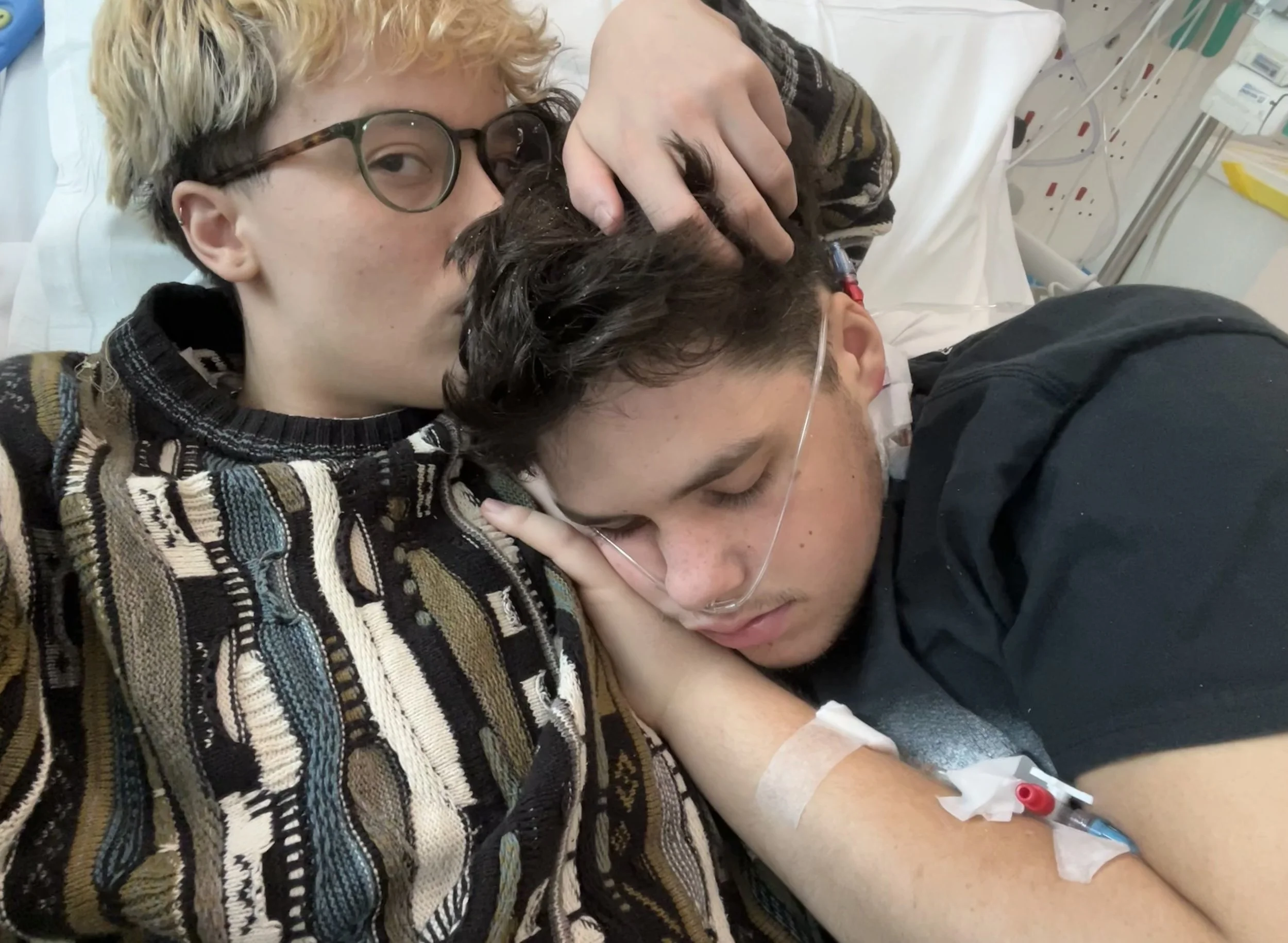 A young person with glasses and blonde hair is lying down in a hospital bed, holding and soothing a patient with dark hair and glasses, who has an IV in their arm and a mask on their face, in a medical setting.