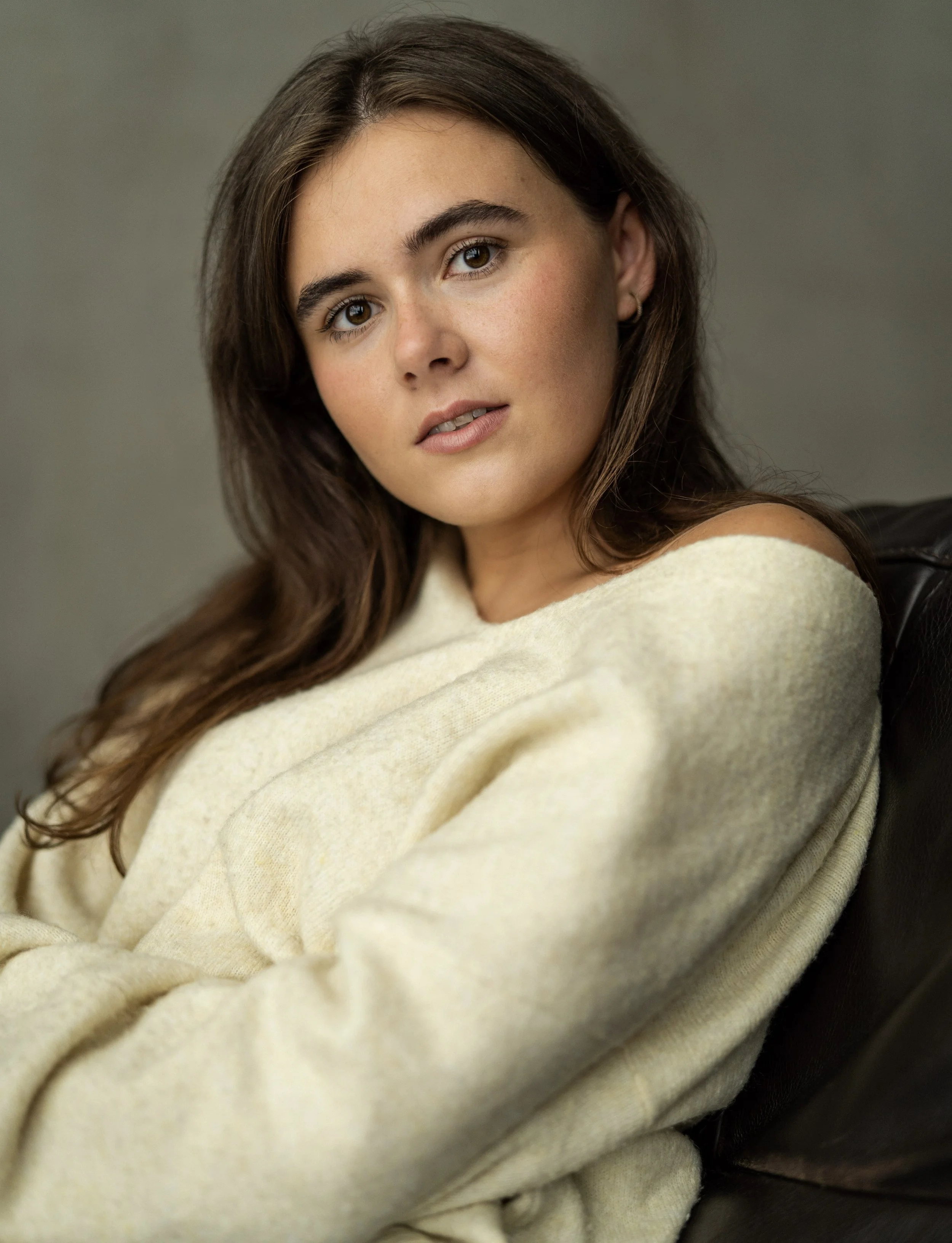 A young woman with long brown hair and brown eyes, wearing a light yellow sweater, is sitting on a dark chair against a plain background, looking softly at the camera.