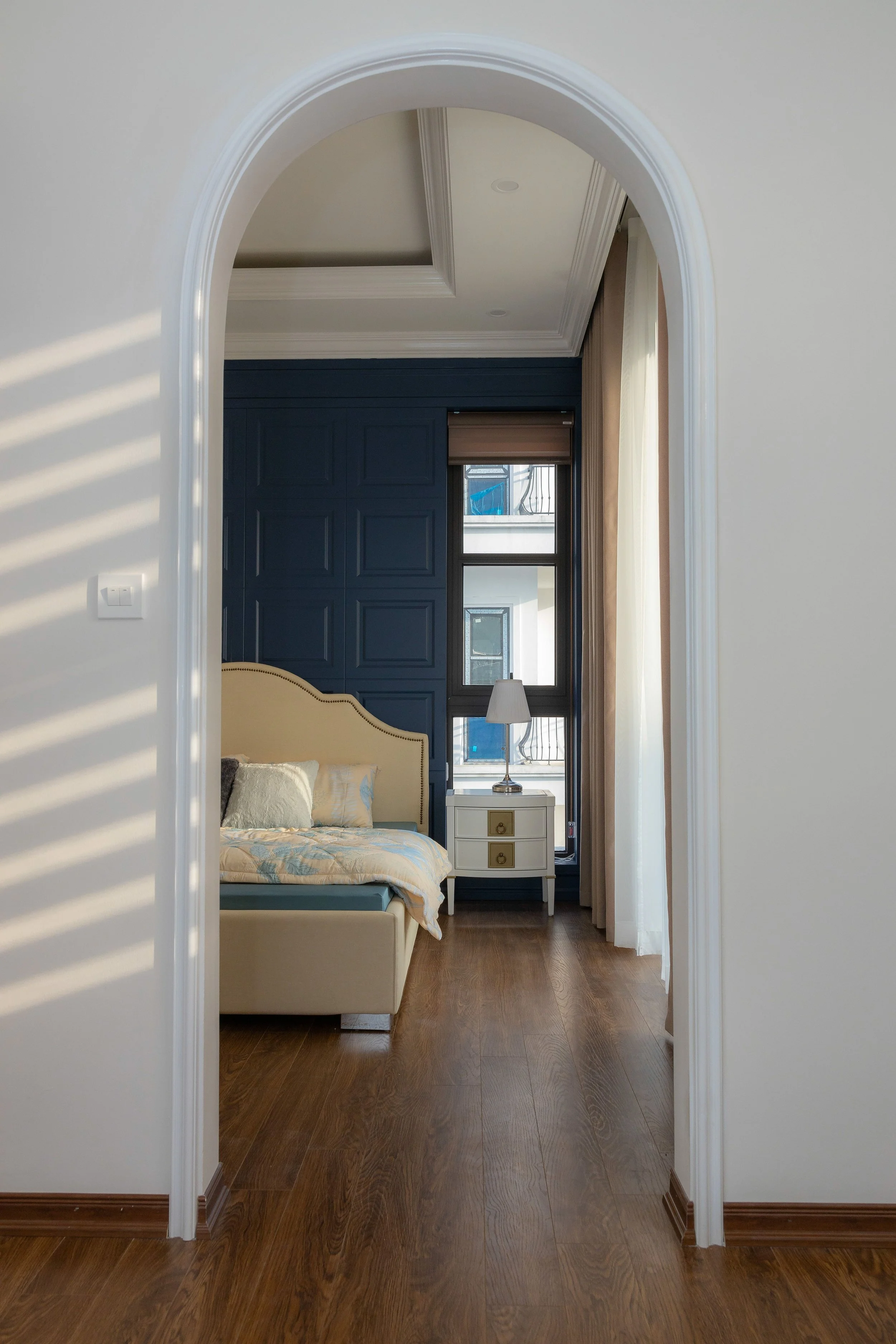 View through an arched doorway into a bedroom with a bed, nightstand, and window with curtains, featuring a blue accent wall.
