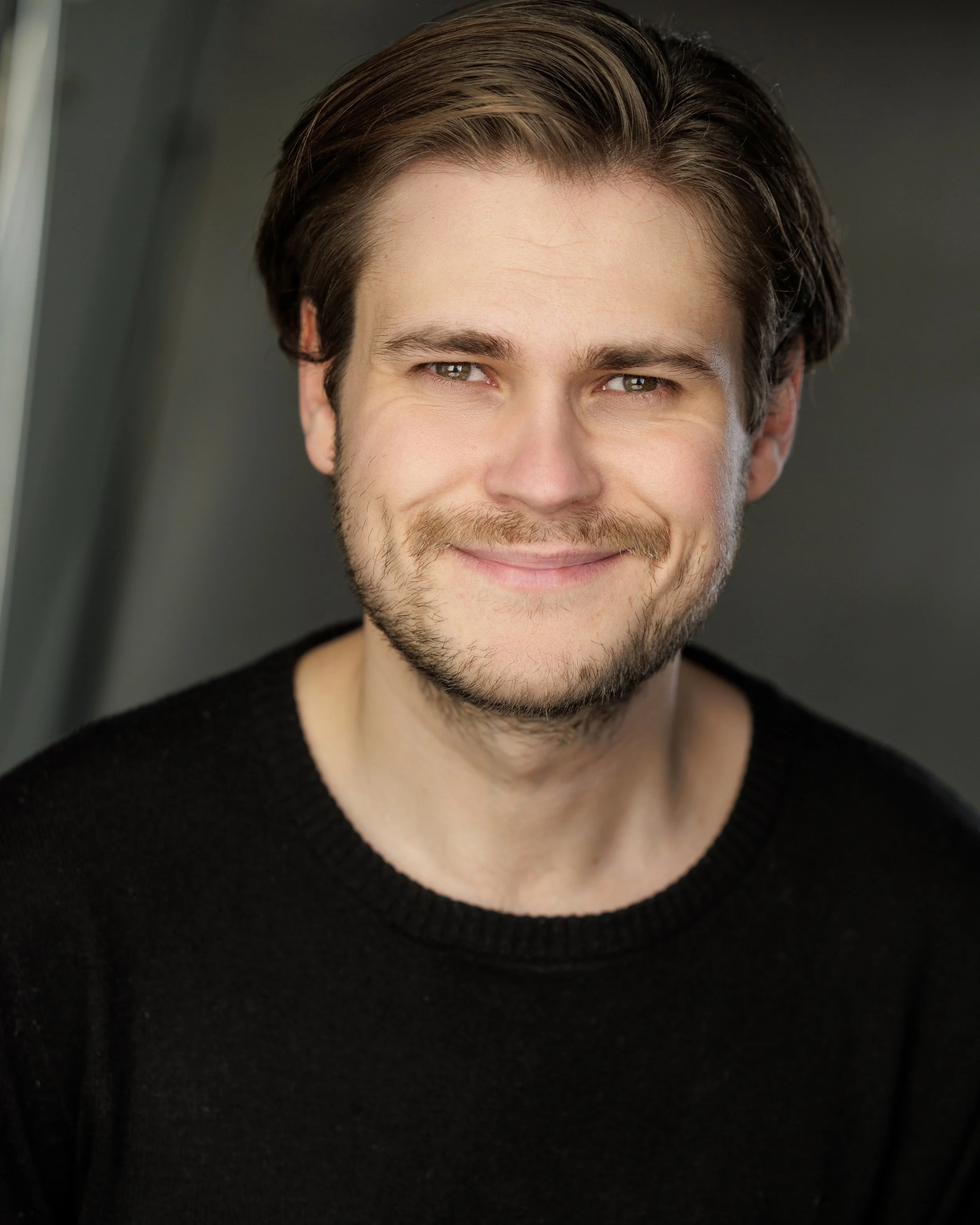 Close-up of a smiling man with brown hair and a beard, wearing a black sweater.