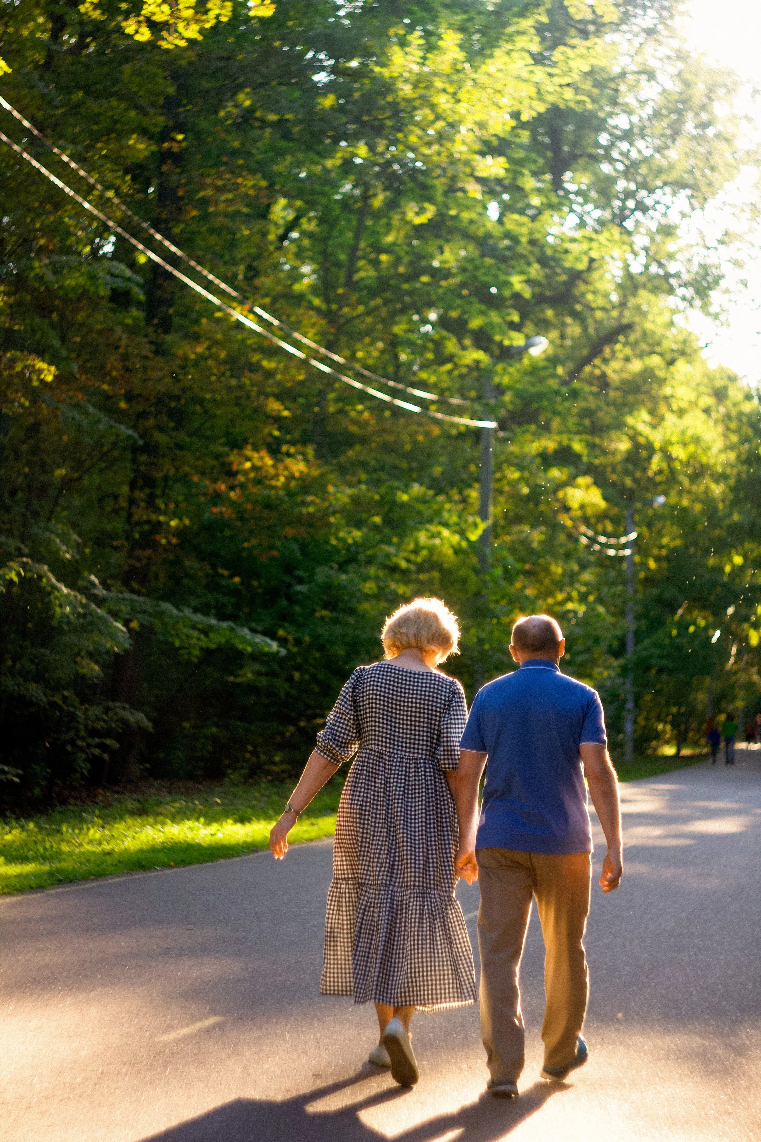 An elderly couple walking hand in hand on a scenic park path during sunset, surrounded by lush green trees with sunlight filtering through.