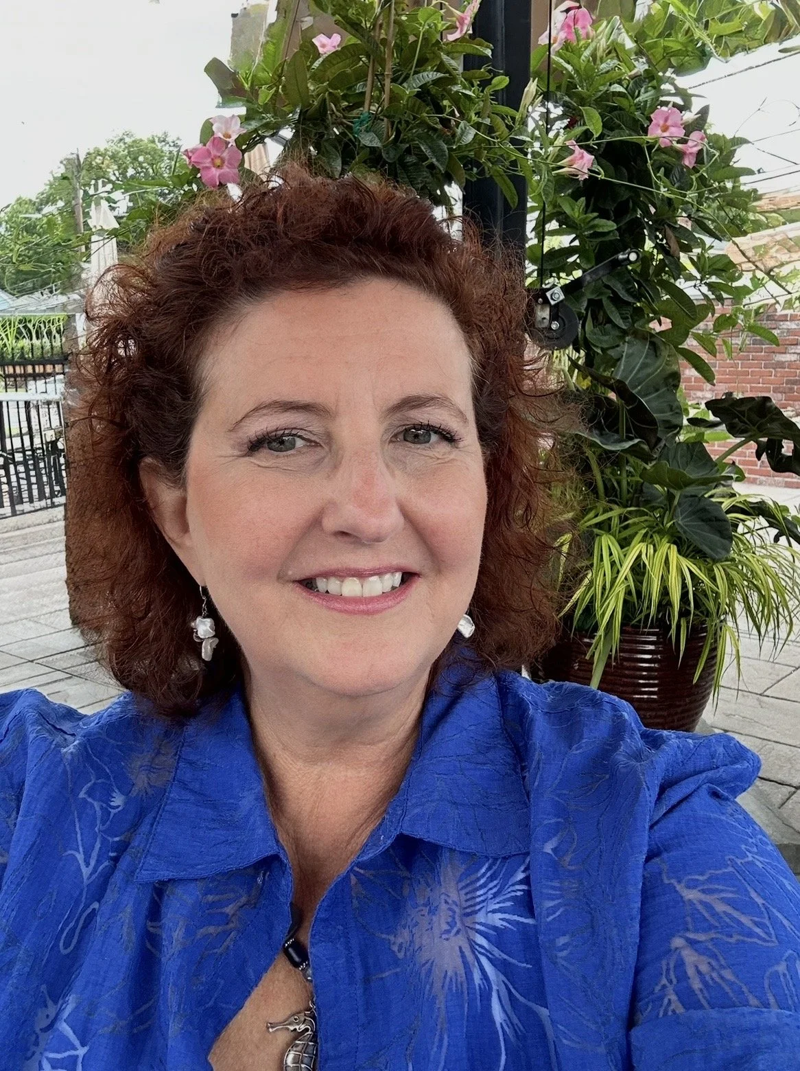 A smiling woman with curly reddish-brown hair, wearing earrings and a blue shirt, taking a selfie outdoors with potted plants and a brick wall in the background.
