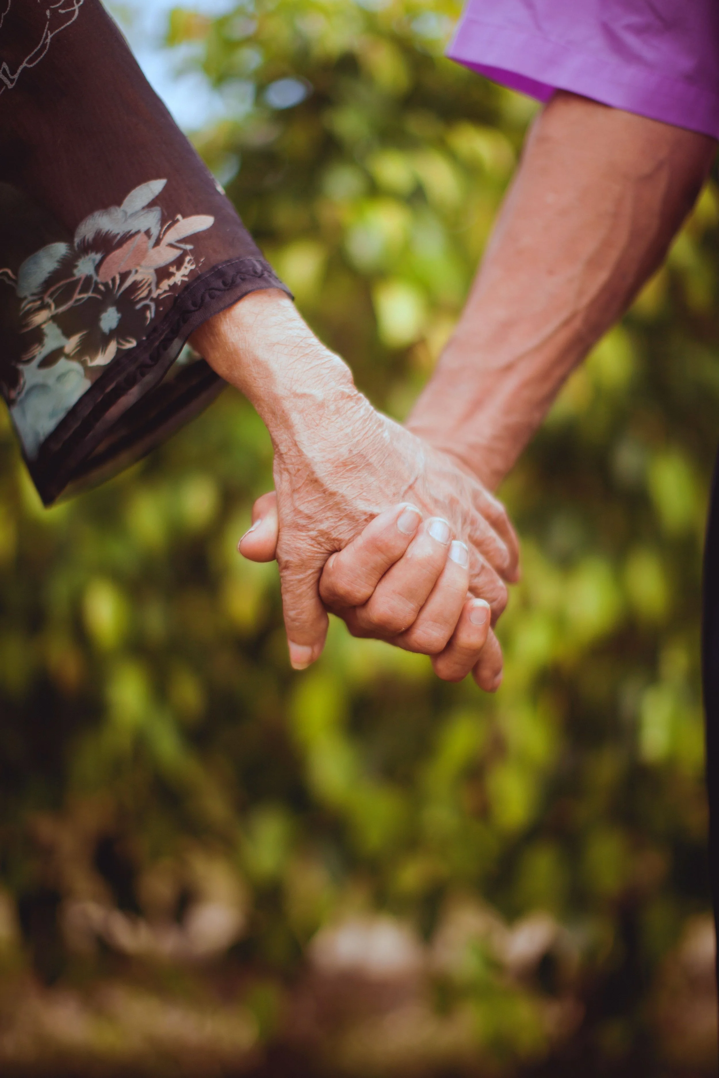 Close-up of elderly hands holding and supporting each other, outdoors with blurred greenery in the background.