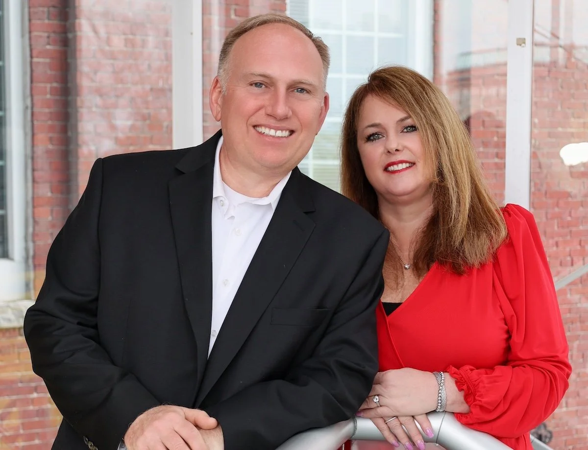 A man and a woman standing together in front of a brick and glass building, both dressed in formal attire, smiling at the camera.