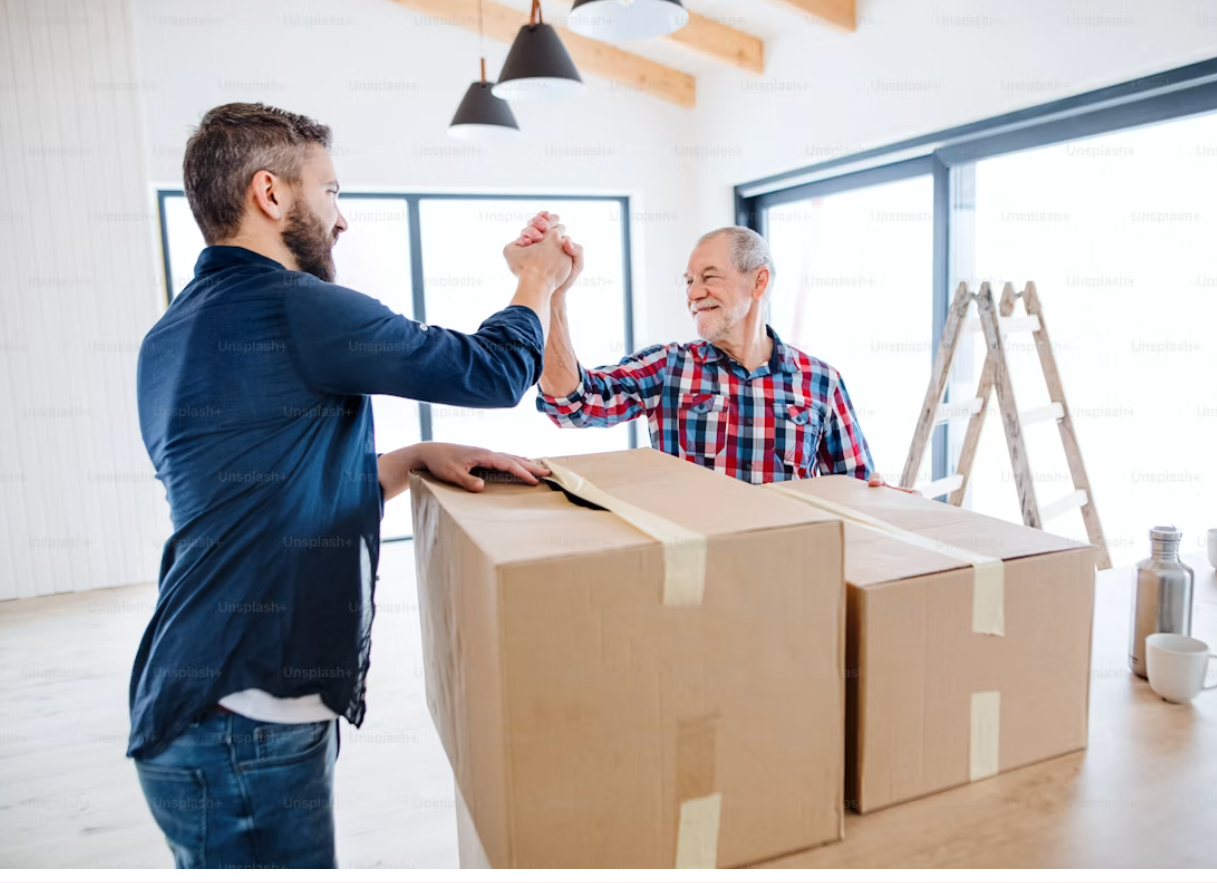 Two men smiling and giving each other an arm wrestle on a large cardboard box in a bright, modern room with large windows, a ladder, and hanging lights.