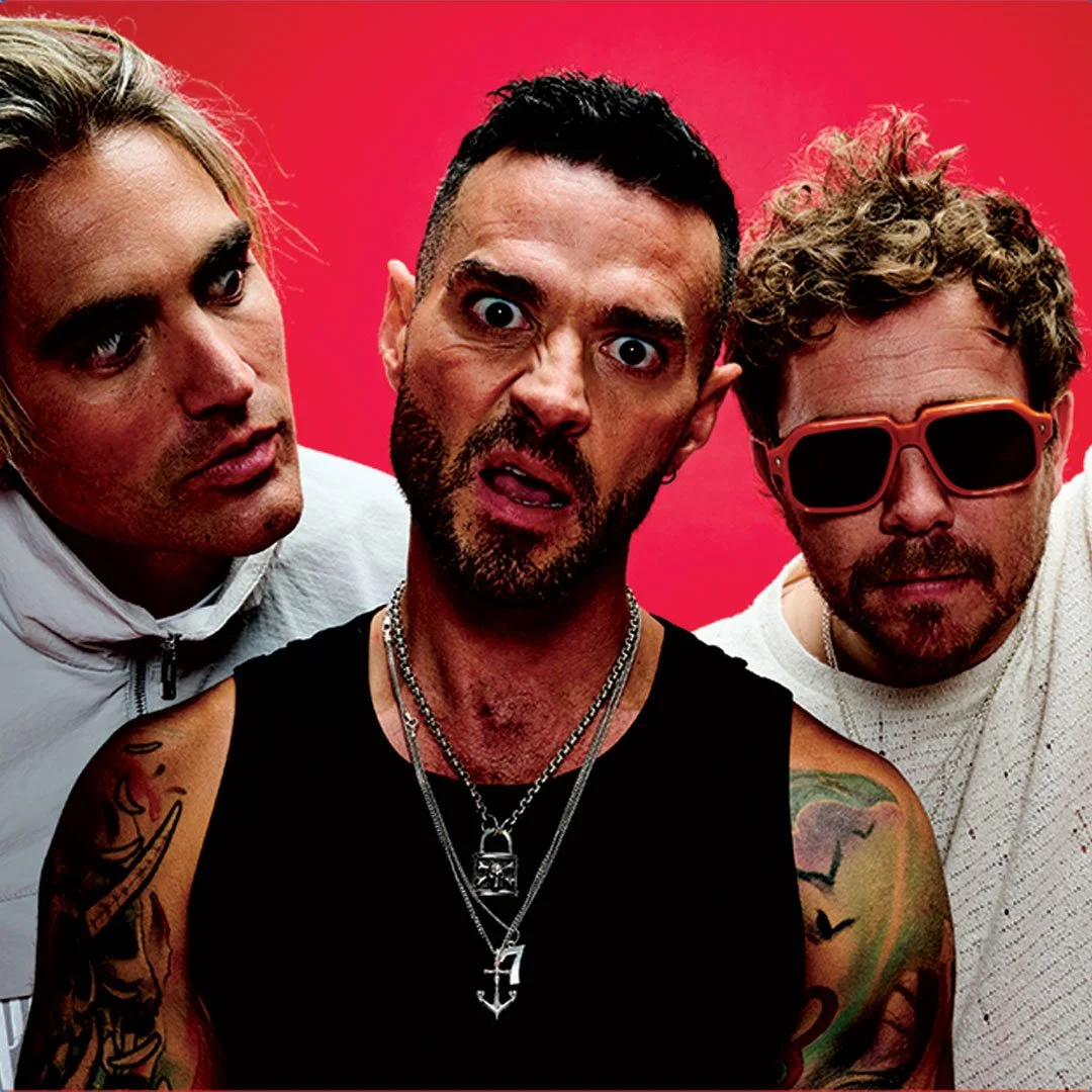 Three men posing together against a red background, one with sunglasses, one with multiple necklaces, and tattoos visible on his arms.